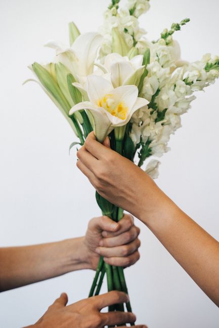 A close-up of a bouquet of fresh flowers being handed over, featuring white lilies with yellow centers, surrounded by white stock flowers, and vibrant green stems. The bouquet is held together with a