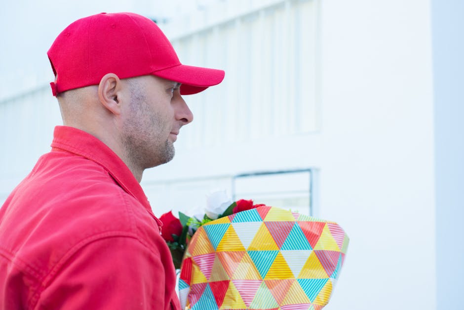A delivery person dressed in a red cap and matching red jacket is holding a vibrant floral gift wrapped in colorful geometric paper with shades of yellow, green, blue, and red. The bouquet features a