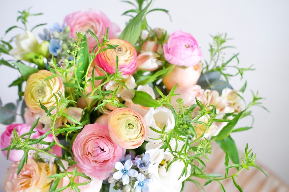 A vibrant floral arrangement featuring pastel pink, cream, and peach ranunculus flowers with tightly layered petals, complemented by small blue and white forget-me-nots. The bouquet includes fresh gre