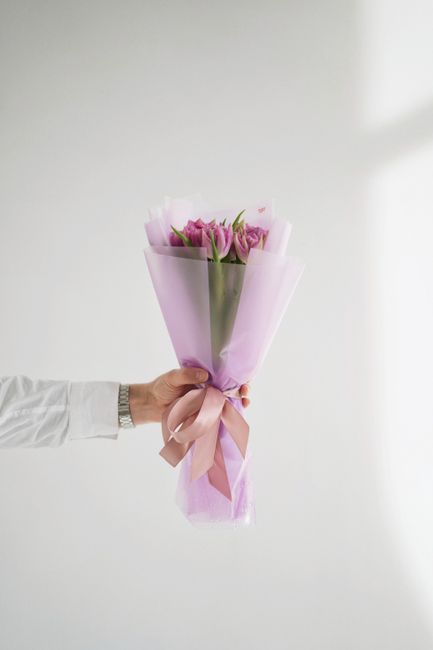 A person wearing a white long-sleeved shirt holds a bouquet of fresh pink tulips, wrapped in light pink tissue paper and tied with a matching pink satin ribbon. The tulips have vibrant green stems and