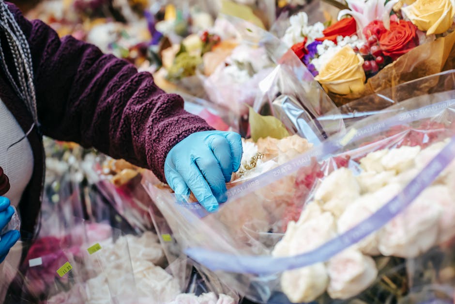 Close-up of a person wearing a purple textured sweater and blue disposable gloves, handling and sorting a bouquet of fresh flowers wrapped in clear plastic. The bouquet contains cream, peach, and purp