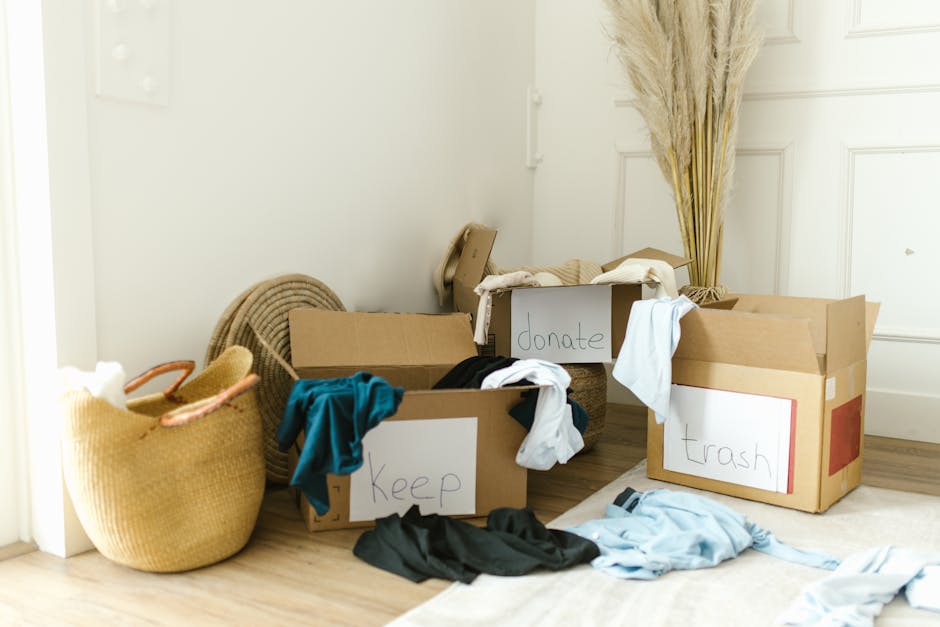 A collection of cardboard boxes on a wooden floor, labeled with handwritten signs reading 'keep', 'donate', and 'trash'. The boxes are filled with various clothing items in different colors, including