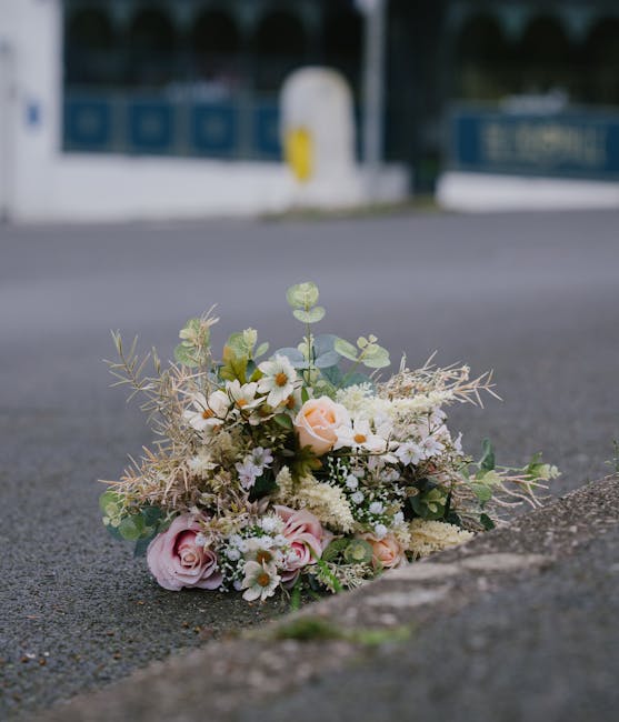 A delicate bouquet of fresh flowers resting on an asphalt street surface near the curb. The arrangement features soft pink and cream roses, white daisies, and tiny white blossoms, complemented by mute