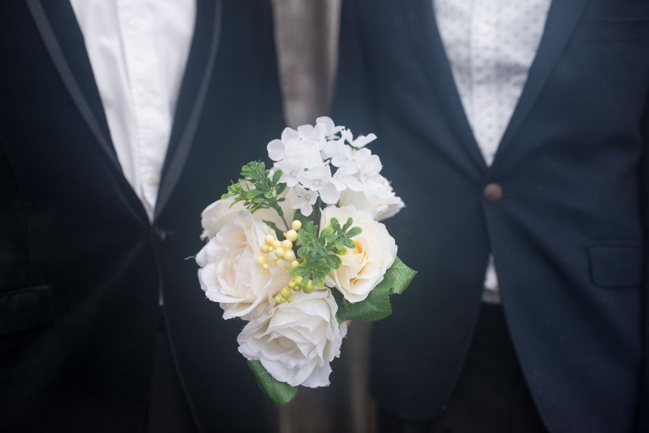 A floral boutonniere featuring white roses, small white hydrangea clusters, and yellow berry accents, accented with green foliage, is being held by a person dressed in a dark formal suit with a light-