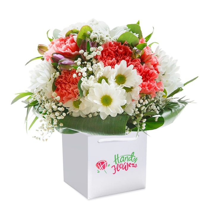 Perfectly poised atop a pure white gift bag, this radiant bouquet seems to echo the urban artistry and welcoming vibrancy of Hoxton itself. The arrangement blends soft coral and peachy red carnations-petals thickly ruffled, hues reminiscent of the evening brickwork along Hoxton Street-with crisp white daisies whose chartreuse centers sparkle brightly. Scattered throughout are tiny, feathery stems of baby's breath, airy and light as the breeze off Gillett Square. Lush leaves and shining green foliage envelop the base, shaping a harmonious cradle that both frames and supports the floral medley. On the left side, subtle purple-tinged blossoms and slender, unopened buds introduce depth and intrigue, contrasting beautifully with the brighter tones. The bouquet's shape is generously rounded, ensuring every stem, from carmine carnation to dainty daisy, is visible and celebrated. The square gift bag-modern with its strong white ribbon handles-offers a practical yet sophisticated delivery solution, a common sight for Hoxton residents surprised by doorstep flowers from loved ones. The backdrop, a blank white canvas, throws every detail into relief, elevating both colour and craftsmanship. This arrangement radiates a sense of celebration, making it a thoughtful gift for birthdays, little victories, or spontaneous acts of kindness, delivered fresh across Hoxton's vibrant streets.